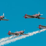 Four vintage propeller planes fly in a tight formation red accents and star insignias leaving white smoke trails in a blue sky