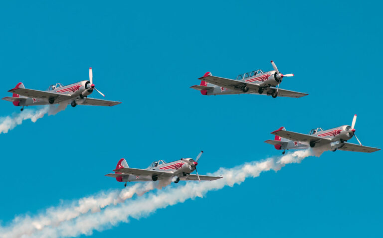 Four vintage propeller planes fly in a tight formation, red accents and star insignias, leaving white smoke trails in a blue sky.