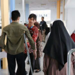 People standing in a bright service hall a man on crutches chats with a man in a red batik shirt while others in hijabs wait nearby near glass doors