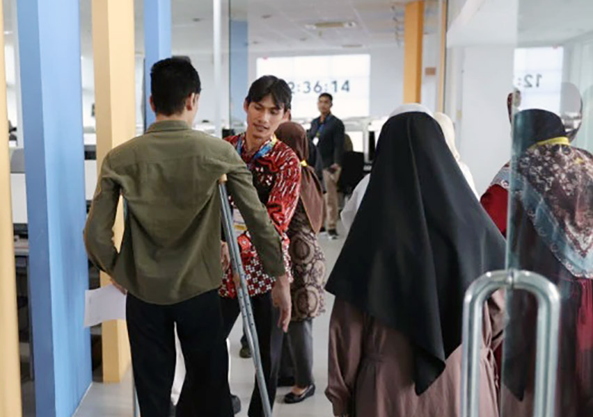 People standing in a bright service hall a man on crutches chats with a man in a red batik shirt while others in hijabs wait nearby near glass doors