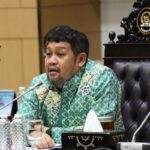 Man in a turquoise batik shirt speaks at a formal meeting seated behind a carved chair with microphones in front of him