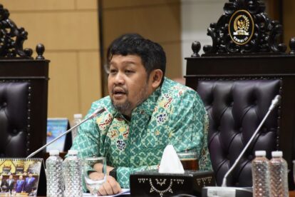 Man in a turquoise batik shirt speaks at a formal meeting seated behind a carved chair with microphones in front of him