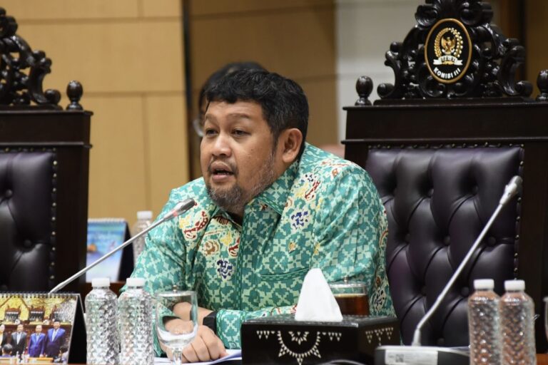 Man in a turquoise batik shirt speaks at a formal meeting, seated behind a carved chair with microphones in front of him.