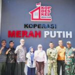 Group photo of men and women standing in front of a blue wall with the sign kop des kel merah putih and a logo outdoors or entrance area