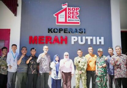 Group photo of men and women standing in front of a blue wall with the sign kop des kel merah putih and a logo outdoors or entrance area