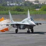 Silver military jet on a tarmac with an orange drag parachute deployed at the rear to aid braking or stopping