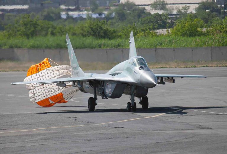 Silver military jet on a tarmac with an orange drag parachute deployed at the rear to aid braking or stopping.