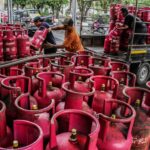 Several workers move and stack red bright gas cylinders on and around a flatbed truck in an urban loading area outside