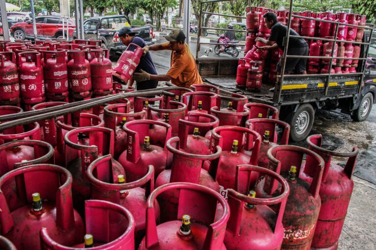 Several workers move and stack red Bright Gas cylinders on and around a flatbed truck in an urban loading area outside.