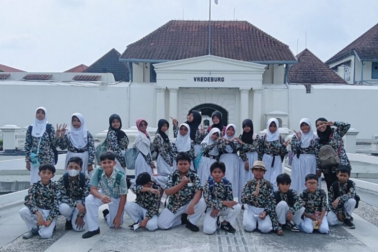 Group of students in matching uniforms posing together on a school trip in front of Vredeburg Fort. The girls wear white hijabs and floral tops, while the boys wear matching shirts and white pants.