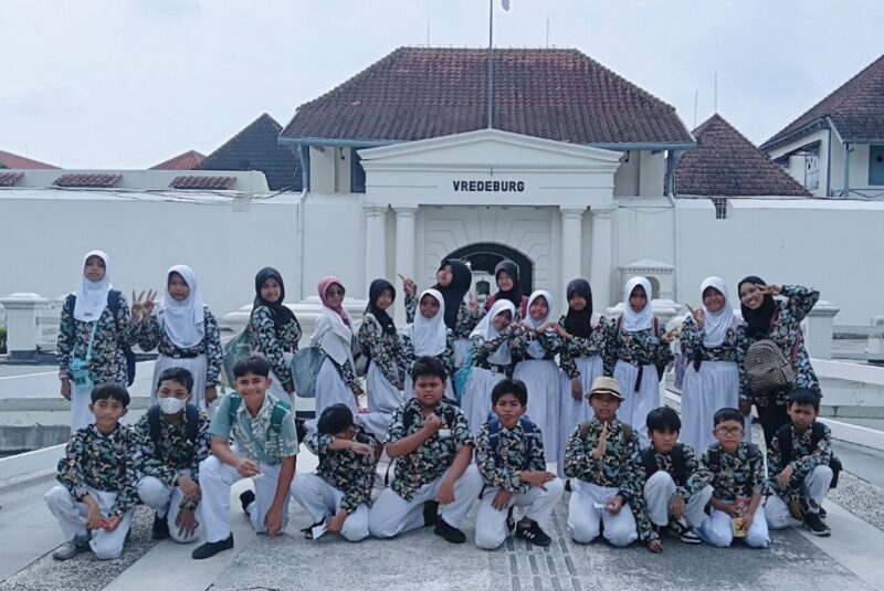 Group of students in matching uniforms posing together on a school trip in front of vredeburg fort The girls wear white hijabs and floral tops while the boys wear matching shirts and white pants