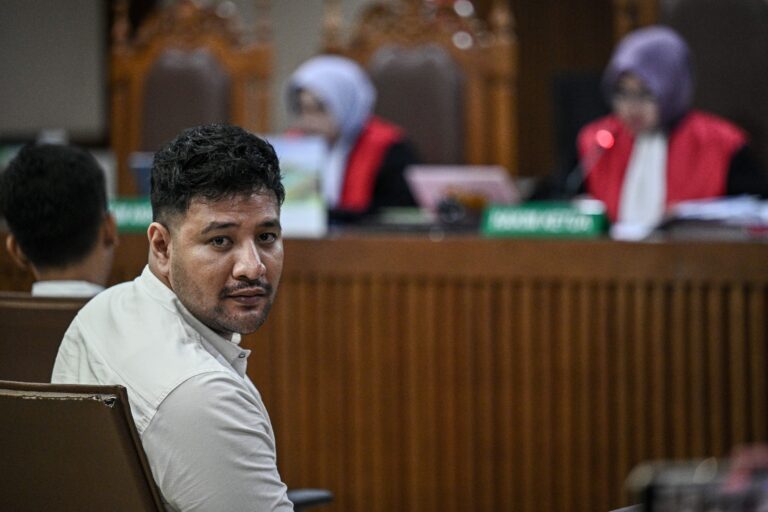 Man seated in a courtroom wearing a light shirt, looking toward the camera with blurred judges in the background.
