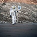 Astronaut in a white space suit standing on a rocky cratered surface while holding a flag with a blue emblem