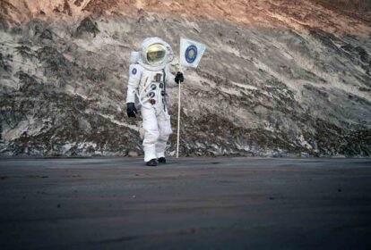 Astronaut in a white space suit standing on a rocky, cratered surface while holding a flag with a blue emblem.