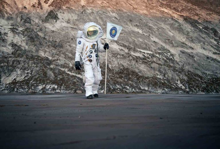 Astronaut in a white space suit standing on a rocky, cratered surface while holding a flag with a blue emblem.