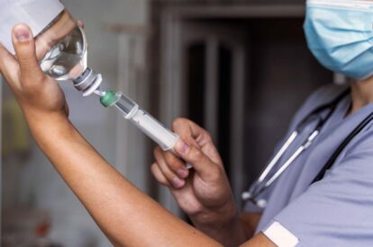 Healthcare professional in scrubs and mask prepares a syringe with a vial in a clinical setting.