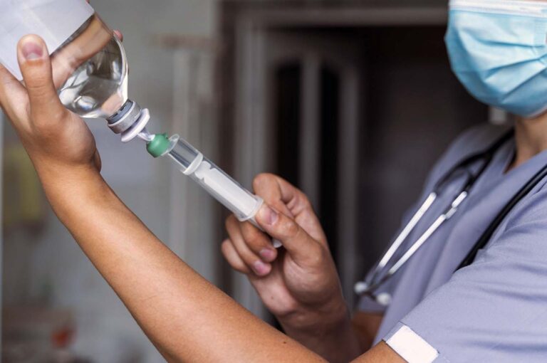 Healthcare professional in scrubs and mask prepares a syringe with a vial in a clinical setting.