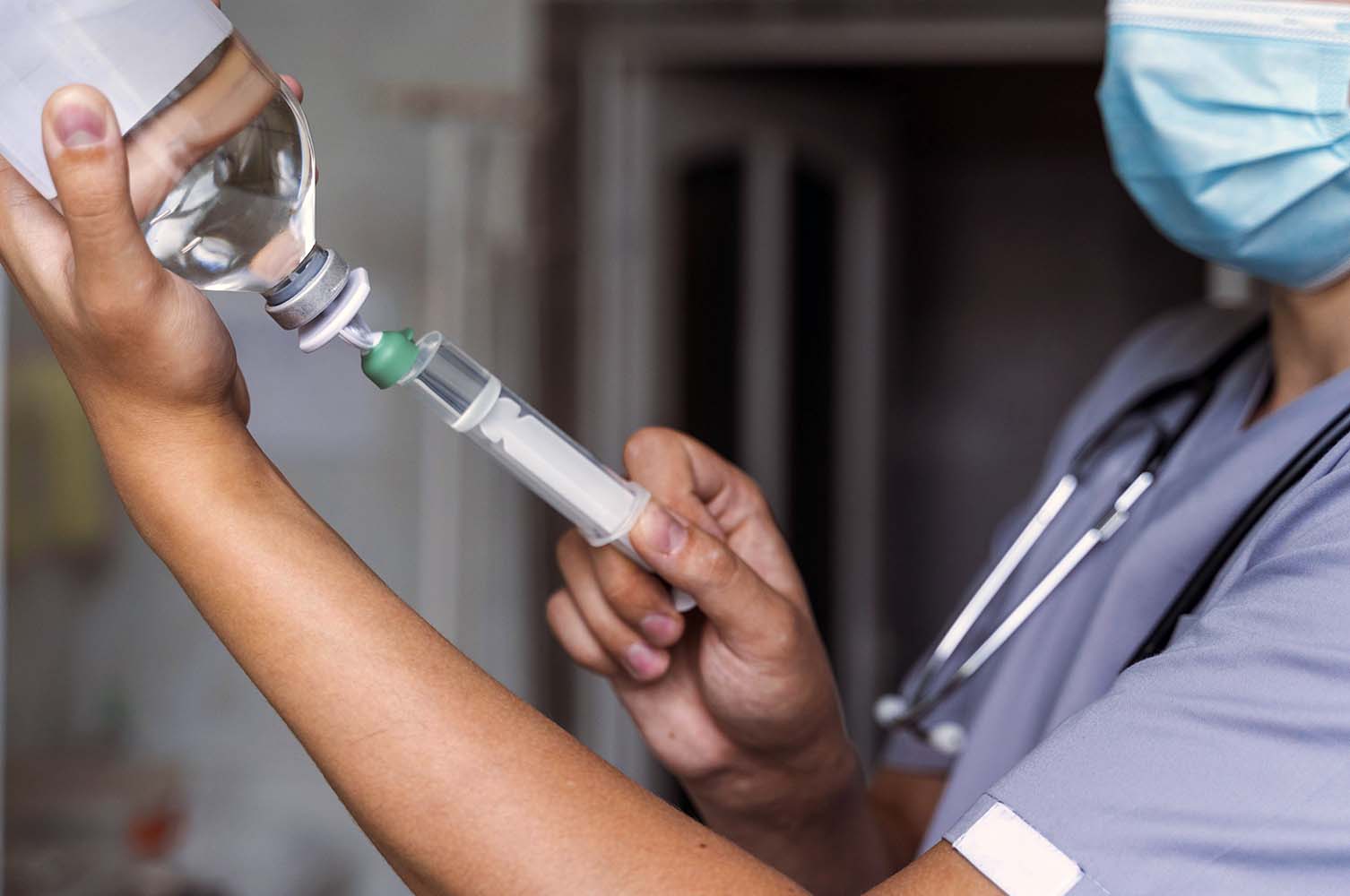 Healthcare professional in scrubs and mask prepares a syringe with a vial in a clinical setting