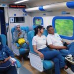People seated in a blue accented train carriage a woman in hijab masks and looks at a phone on the left others sit by windows with greenery visible outside