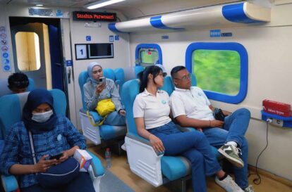 People seated in a blue-accented train carriage; a woman in hijab masks and looks at a phone on the left, others sit by windows with greenery visible outside.