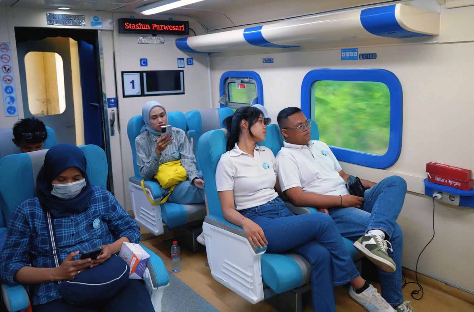 People seated in a blue accented train carriage a woman in hijab masks and looks at a phone on the left others sit by windows with greenery visible outside
