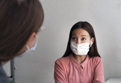 Two people wearing face masks converse in an indoor setting woman in a pink blouse looking surprised or engaged at the other person