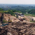 Open pit quarry with excavator and dump trucks on a dirt road brown rocky piles and green hills in the background