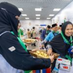 Smiling woman in a black hijab hands a wicker basket of pastries to a guest at an indoor event