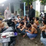 Police officer standing among a group of shirtless men seated on the ground outside a house motorcycles parked nearby
