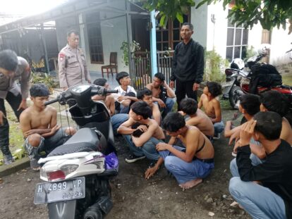 Police officer standing among a group of shirtless men seated on the ground outside a house motorcycles parked nearby