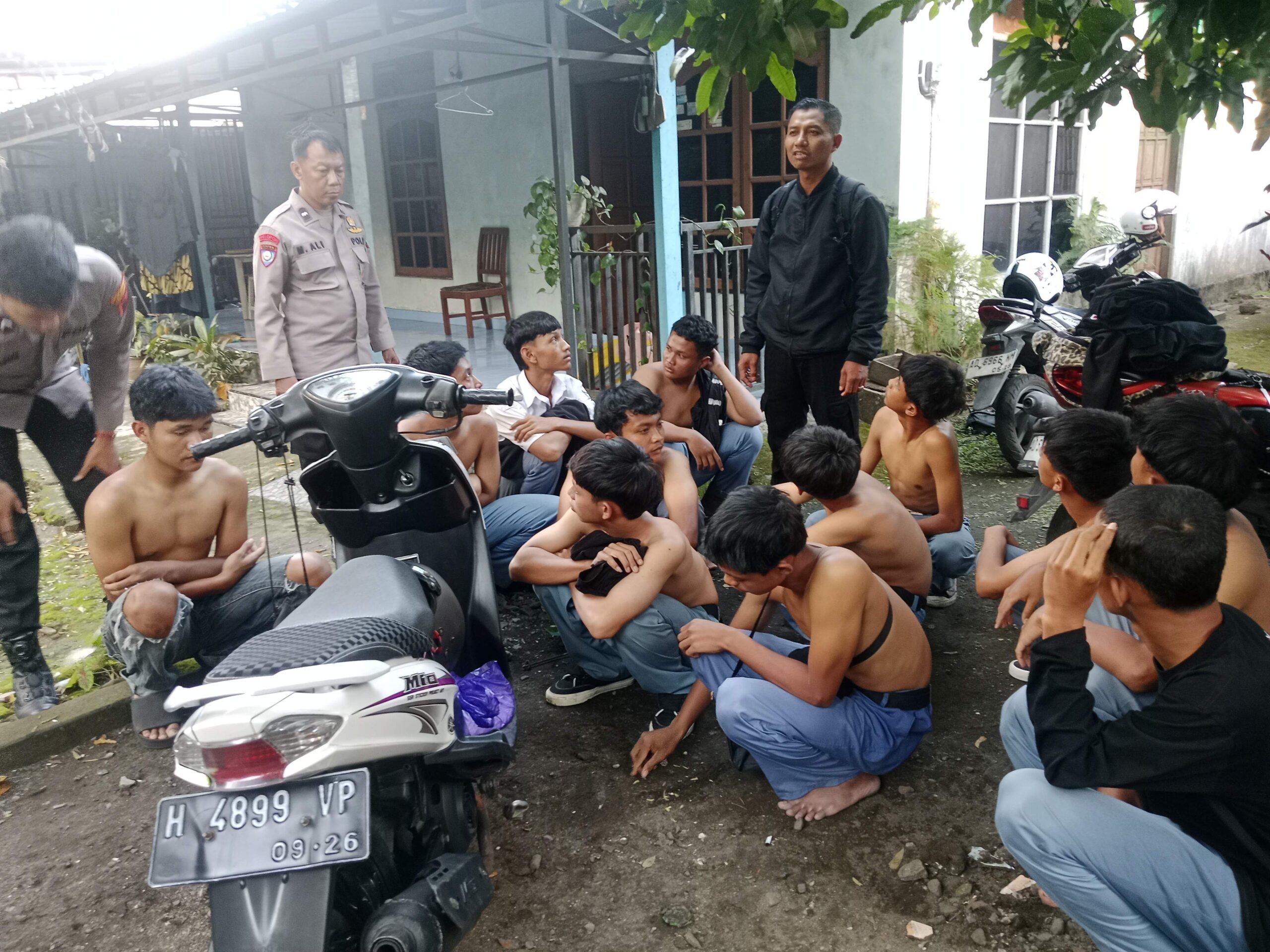 Police officer standing among a group of shirtless men seated on the ground outside a house motorcycles parked nearby