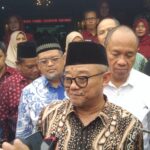 Central man in batik shirt and black peci speaks to reporters as microphones are held up crowd of people behind him