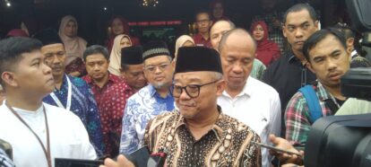 Central man in batik shirt and black peci speaks to reporters as microphones are held up; crowd of people behind him.