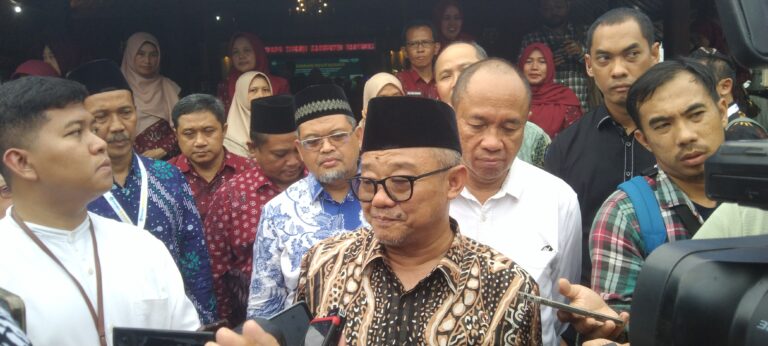 Central man in batik shirt and black peci speaks to reporters as microphones are held up; crowd of people behind him.