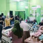 Busy hospital ward with green walls patients on beds connected to ivs surrounded by family and staff