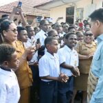 A man in a light blue shirt shakes hands with a group of students and officials as others take photos in a crowded outdoor courtyard
