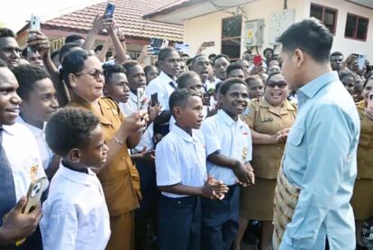 A man in a light blue shirt shakes hands with a group of students and officials as others take photos in a crowded outdoor courtyard.