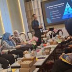 Presenter in a dark suit explains a plan at the front of a conference room while attendees in hijabs sit around a long table with papers water bottles and pink orchids a projection screen shows a blue pyramid diagram labeled in indonesian