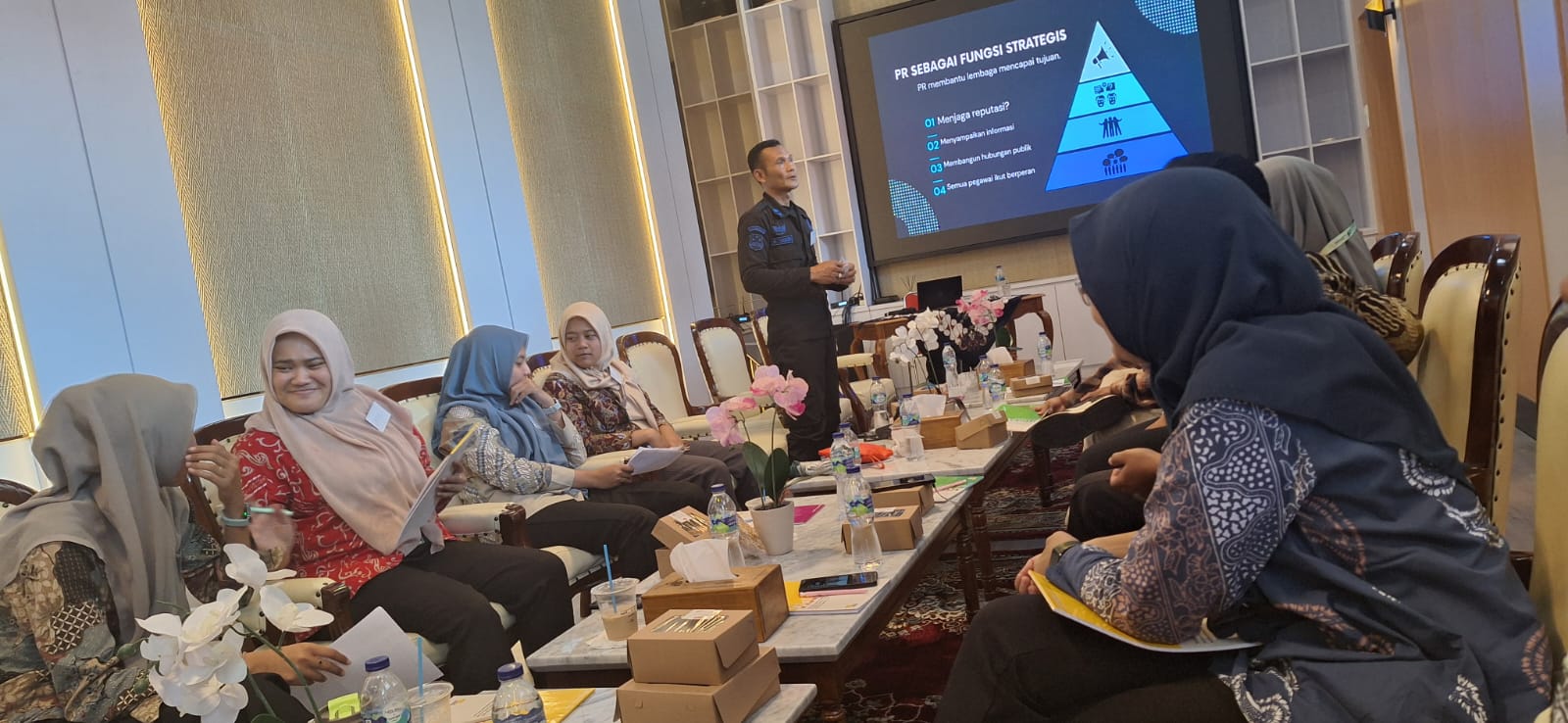 25latih catrawarta Presenter in a dark suit explains a plan at the front of a conference room while attendees in hijabs sit around a long table with papers water bottles and pink orchids a projection screen shows a blue pyramid diagram labeled in indonesian