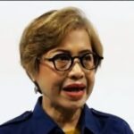 Woman with short blonde hair and large black glasses speaking wearing a navy blue shirt against a light background