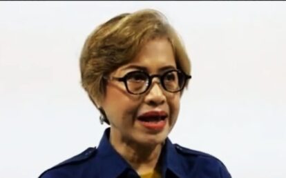 Woman with short blonde hair and large black glasses speaking, wearing a navy blue shirt against a light background.
