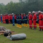 Line of emergency responders in red and blue uniforms and helmets standing at attention on a grassy field with equipment laid out in front
