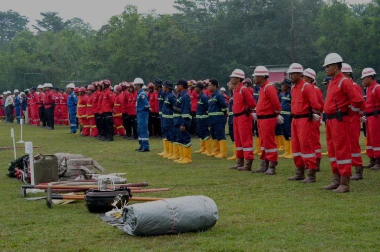 Line of emergency responders in red and blue uniforms and helmets standing at attention on a grassy field with equipment laid out in front.