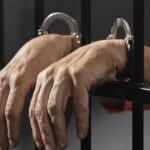 Close up of a persons hands in metal handcuffs resting on jail bars conveying detention