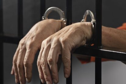 Close-up of a person’s hands in metal handcuffs resting on jail bars, conveying detention.
