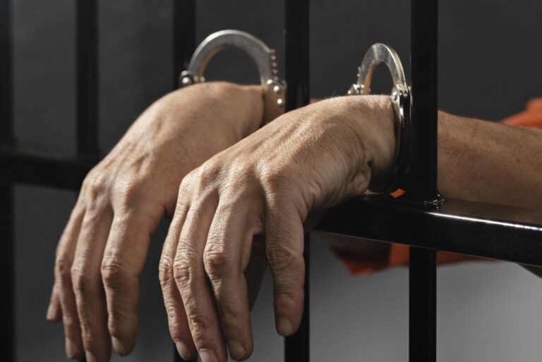 Close-up of a person’s hands in metal handcuffs resting on jail bars, conveying detention.