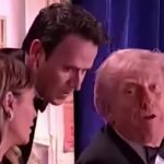 Old man in a dark tuxedo looks toward the camera while two people stand beside him near a blue curtain