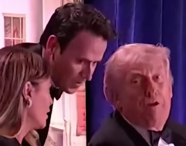 Old man in a dark tuxedo looks toward the camera while two people stand beside him near a blue curtain.