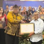 Man in yellow batik hands a certificate to a smiling man in a white shirt during an award ceremony group of colleagues in background holding plaques