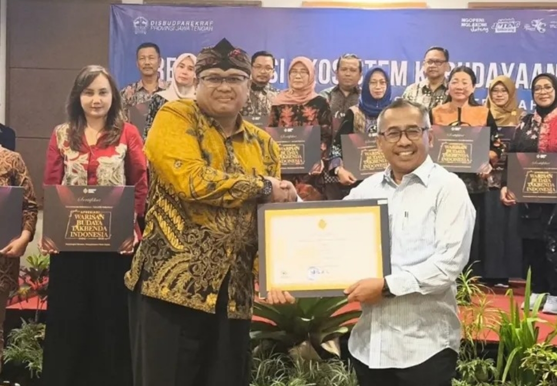 26ilir catrawarta Man in yellow batik hands a certificate to a smiling man in a white shirt during an award ceremony group of colleagues in background holding plaques